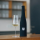 A black fabric sleeve has been slipped over a wine bottle, which stands next to a filled wine glass on a wooden table.